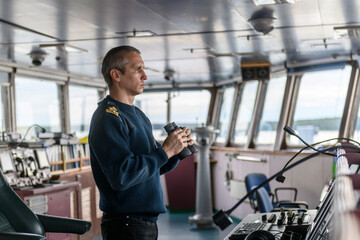 Deck officer with binoculars on navigational bridge. Seaman on board of vessel. Commercial shipping. Cargo ship. - obrazy, fototapety, plakaty