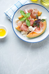 Cantaloupe slices with prosciutto, torn mozzarella and fresh basil served in a plate, flat lay on a light-grey granite background, vertical shot, copyspace