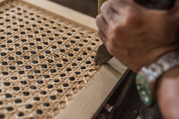 A carpenter removes staples securing a rattan solihiya screen from a wooden frame with a pair of pliers. A rattan furniture workshop.