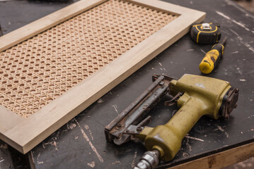A cabinet panel with a rattan solihiya weave pattern on a workshop table beside a pneumatic staple...