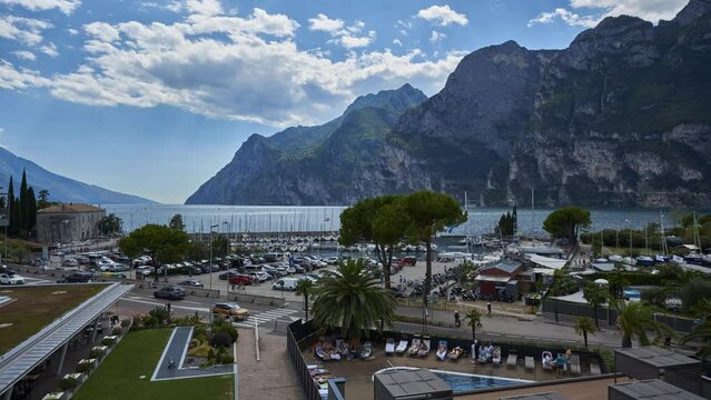 TimeLapse of the port area of Riva del Garda and the clouds moving over the lake and the mountains. Transport of vehicles along the lakefront and tourists who enjoy the services of the place.