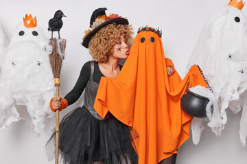 Female witch in black dress embraces and wants to kiss ghost holds broom with crow expresses love to creepy creatures poses against decorated background believes in mystery happen on Halloween night