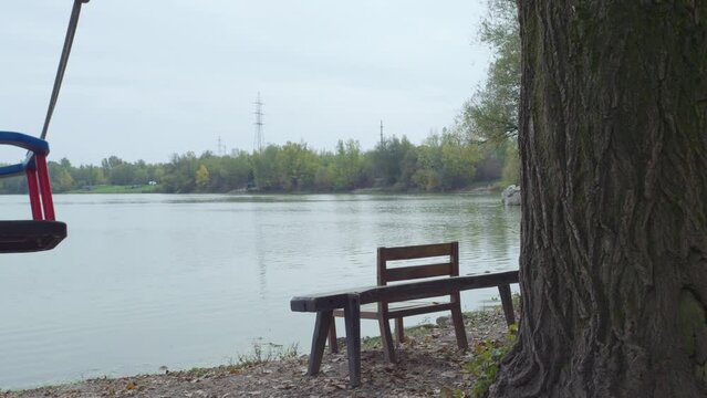 Panning footage of the empty wooden table, chairs, and sway swaying by the lake shore