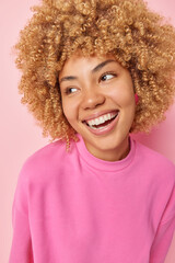 Vertical shot of happy woman with fair curly hair smiles broadly shows white teeth focused aside being in good mood has carefree expression wears casual pullover poses indoors. Emotions concept