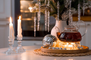 A bouquet of fir branches with icicles, candles and a teapot with tea on a wooden table in the home interior of a white Scandinavian-style kitchen. Cozy concept of festive drinks.