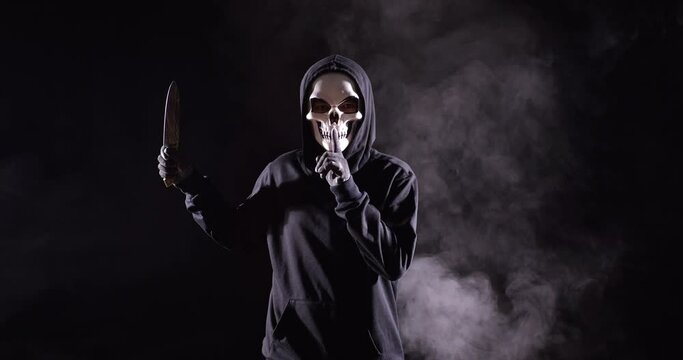 Scary Man In The Hooded Sweatshirt Wearing Halloween Mask Gesturing Silence While Holding A Knife On The Black Background With Smoke
