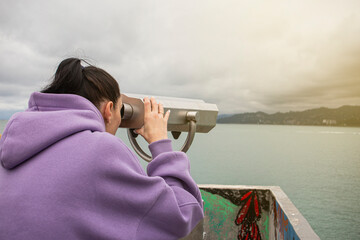 a girl watching through stationary binoculars on the pier for passing ships and wildlife on the...