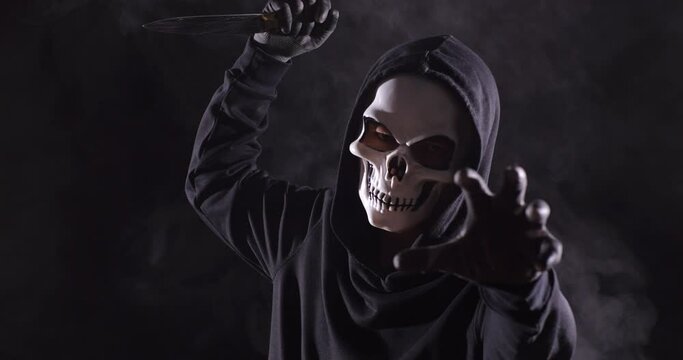 Close Up Of Scary Man In The Hooded Sweatshirt Wearing Halloween Mask Holding A Knife In The Threatening Gesture, A Symbol Of Murder And Crime, On The Black Background With Smoke
