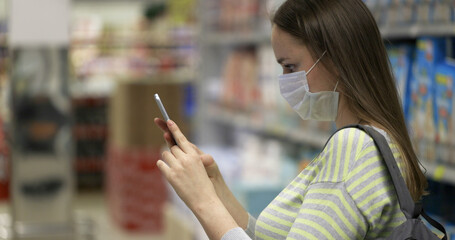The buyer takes pictures on a smartphone in the store. A woman in a yellow and gray sweater and a protective mask makes purchases in the supermarket. Daily lifestyle. 