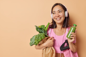 Positive Asian woman carries paper bag with vegetables holds bottle of green fresh smoothie keeps...