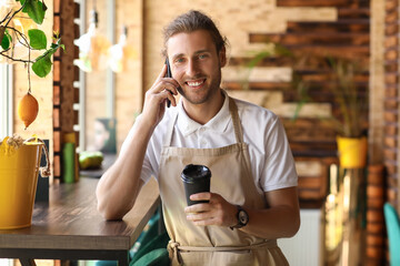 Handsome business owner with coffee cup talking by mobile phone in his cafe