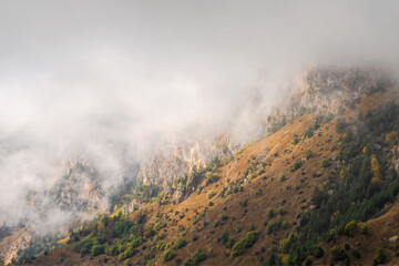 Mountains in a dense fog and sunny slope. Mystical landscape with beautiful sharp rocks in low clouds. Beautiful mountain foggy scenery on abyss edge with sharp stones.