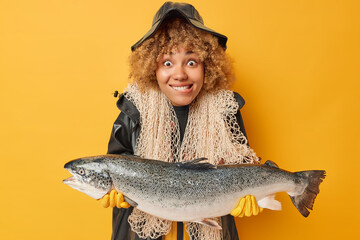 Positive fisherwoman with curly hair bites lips holds huge caught salmon carries fishnet around...