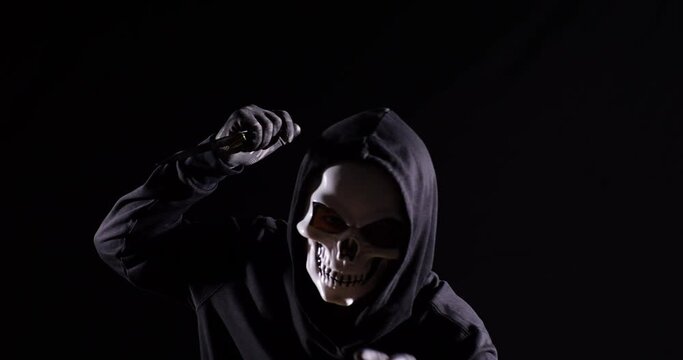 Close Up Of Scary Man In The Hooded Sweatshirt Wearing Halloween Mask Holding A Knife And Making Frightening Gesture While Running On The Black Background
