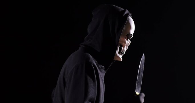 Close Up Side View Of Scary Man In The Hooded Sweatshirt Wearing Halloween Mask Holding A Knife And Running On The Black Background 
