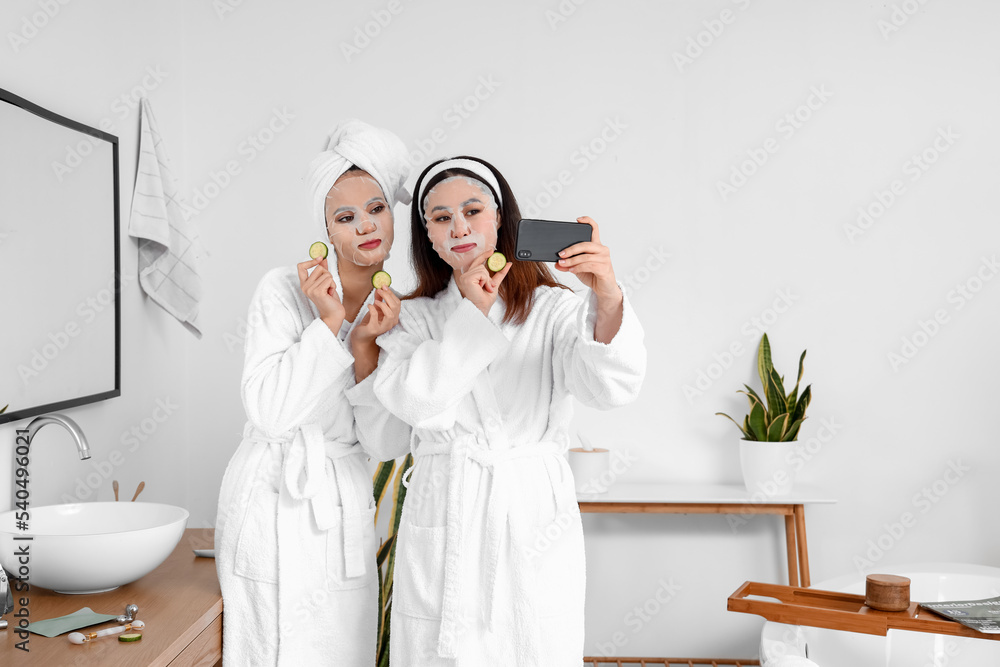 Wall mural Beautiful young women with sheet facial masks and cucumber slices taking selfie in bathroom