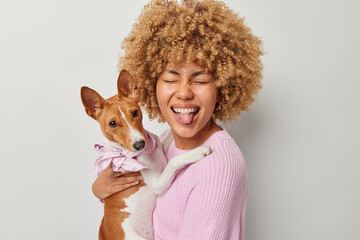Happy joyful woman with curly hair sticks out tongue foolishes around holds pedigree dog going to have walk dressed in pink knitted jumper poses indoor. Display of affection. Friendship concept