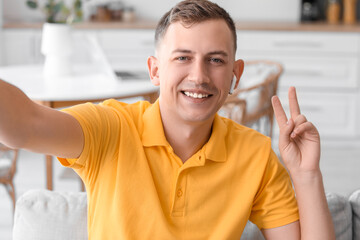 Young man showing victory gesture while taking selfie on sofa at home