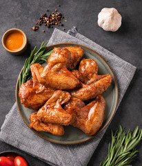 Spicy grilled chicken wings with sauce on a plate with tomato and herb on a dark background of slate or concrete.