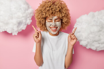 Waist up shot of positive young woman with curly hair applies beauty mask for reducing fine lines points index fingers above demonstrates somemthing isolated over pink background clouds overhead