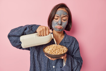 Serious brunette Asian woman pours milk in cornflakes applies beauty mask to reduce pores dressed...