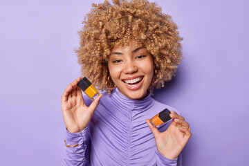 Portrait of good looking woman with curly hair holds two bottles of nail polish smiles positively wears golden earrings and turtleneck isolated over purple background. Beauty treatment and hand care