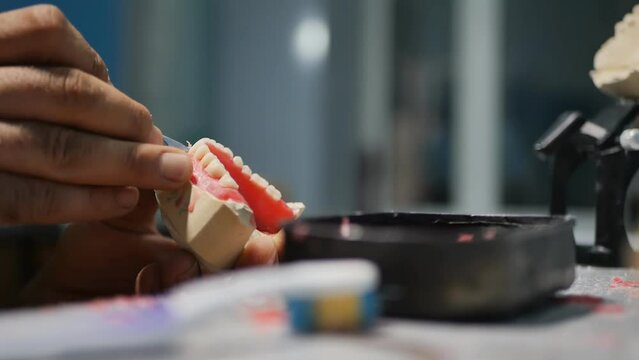 The hands of a dental technician make an artificial replaceable denture for a client holding the denture in his hands and cutting the paraffin from which the gums are made with a sharp scalpel