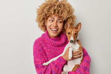 Photo of cheerful European woman embraces own pet enjoys company of pedigree dog wears knitted...