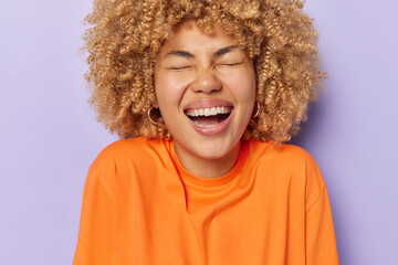 Cheerful young woman with curly bushy hair laughs happily keeps eyes closed expresses positive...