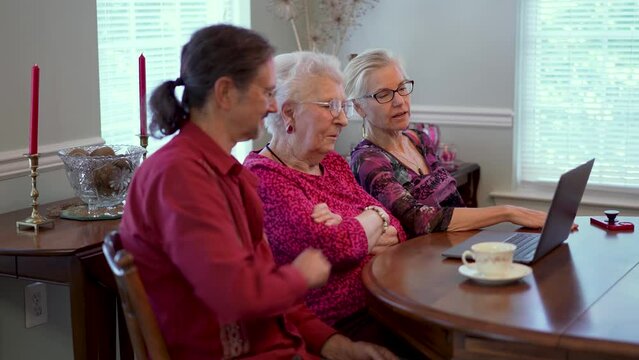 Closeup of elderly senior mother with mature son and daughter in law in dining room sitting in front of laptop computer looking at photos and talking about memories.