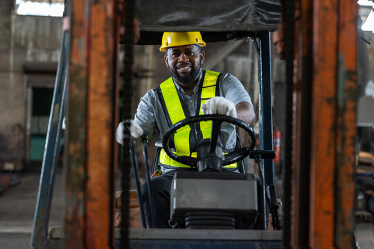 African American male worker driving forklift truck in heavy metal industrial factory. Smiling man engineer wearing vest and helmet safety working and moving heavy metal material at warehouse factory. - Powered by Adobe