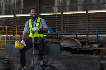 African American male worker wearing vest and helmet safety sitting relaxed after working by using shovel to scoop coal on floor for burning at heavy metal industrial factory
