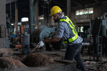 African American male worker wearing vest and helmet safety using shovel to scoop coal on floor for burning at metal factory. Man technician foreman working heavy metal machine at industrial factory. - Powered by Adobe