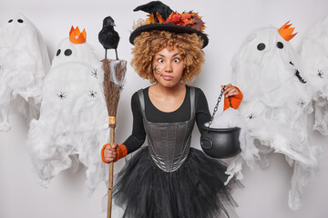 Funny curly haired female wizard with spider on face crosses eyes makes grimace holds broom and cauldron involved in witchcraft wears black hat dress poses around spooky ghosts. Halloween is coming - obrazy, fototapety, plakaty