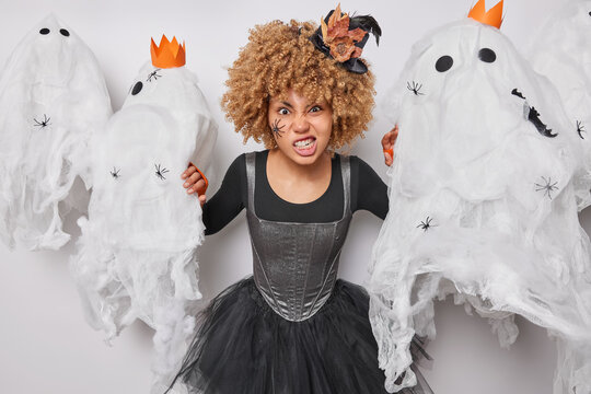 Photo of irritated woman with curly hair clenches teeth and looks angrily at camera wears black dress and hat prepares for Halloween holiday surrounded by scarying creatures. Autumn holiday concept