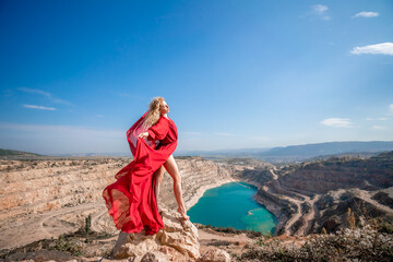 Side view of a beautiful sensual woman in a red long dress posing on a rock high above the lake in the afternoon. Against the background of the blue sky and the lake in the form of a heart