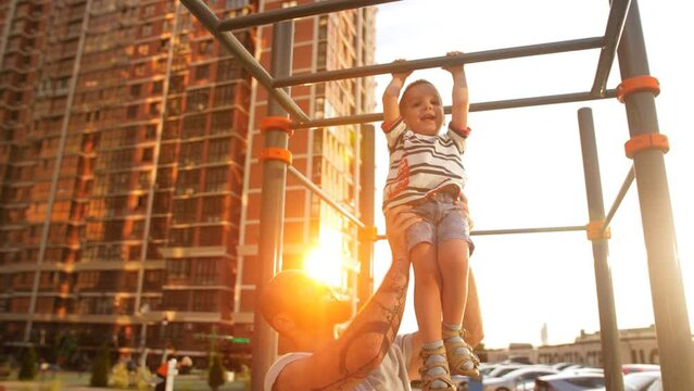 Father supporting little son while she going across monkey bars on outdoor workout playground on sunny day.