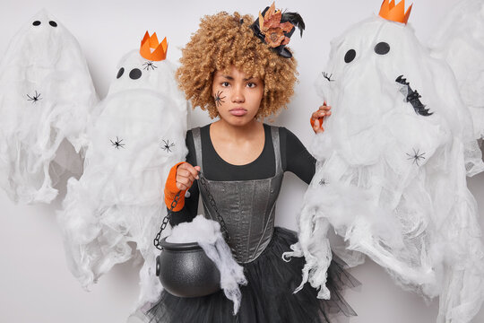 Sad woman dresses up costume holds cauldron tries to be scary involved in witchcraft looks upset stands around creepy creatures against white background. Halloween celebration on 31st of October