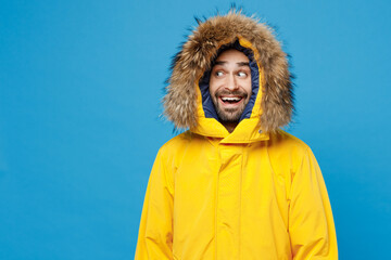 Young smiling fun man 20s wearing yellow puffer down jacket with fur hood look aside on workspace area mock up isolated on plain blue color background studio portrait. People winter lifestyle concept. © ViDi Studio