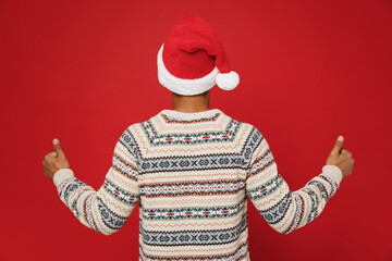 Fototapeta na wymiar Back view merry young man wear cozy Christmas sweater Santa hat posing showing thumb up like gesture wink blink eye isolated on plain red background. Happy New Year 2023 celebration holiday concept.