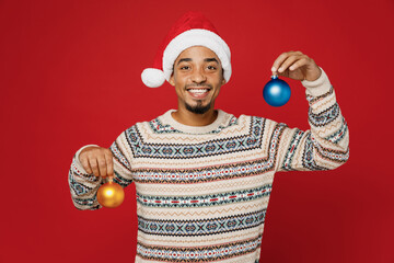 Merry smiling young man wear warm cozy sweater Santa hat posing hold in hand two toys for Christmas tree decorations isolated on plain red background. Happy New Year 2023 celebration holiday concept.