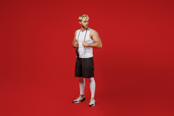 Fototapeta na wymiar Full body young strong sporty sportsman man wears white clothes spend time in home gym stand with skipping rope over neck look aside isolated on plain red background. Workout sport fit body concept.