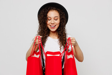 Young fun smiling happy woman wearing t-shirt hat hold in hand look open red paper package bags after shopping isolated on plain solid white color background studio. Black Friday sale buy day concept.