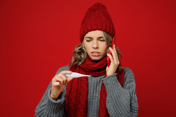 Young woman wear grey sweater scarf hat talk speak on mobile cell phone coucgh hold thermometer isolated on plain red background. Healthy lifestyle ill sick bad disease treatment cold season concept.