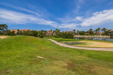 Fototapeta na wymiar Beautiful view of Aruba's hotel golf course. Green grass and pink buildings behind Green trees on blue sky with white clouds background. 