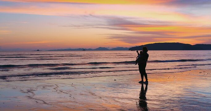 Silhouette of happy family mother  playing carrying cute baby daughter at the beach during amazing sunset enjoying their vacation together at ao nang beach, krabi Thailand