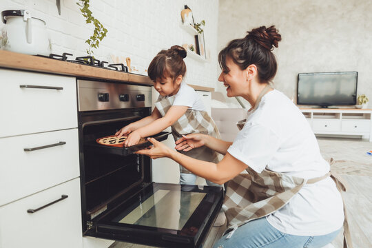 A little girl and her mom put a baking dish with a pie in the oven in the kitchen. mom and child cook a pie together in the kitchen.