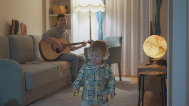 Happy young father and his son fun in cozy home living room. Lonely dad read play guitar for his kid. Funny boy dancing. Multiracial family. Parenthood childhood concept, daddy in maternity leave.