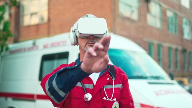 Close up of senior Caucasian professional paramedic in red uniform wearing VR headset and making gestures with hands in air standing outdoors. Medical worker. Futuristic. Virtual reality, metaverse