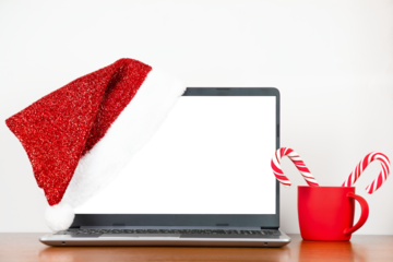 Santa hat on laptop with blank monitor, red cup with candy canes on wooden table, copy space